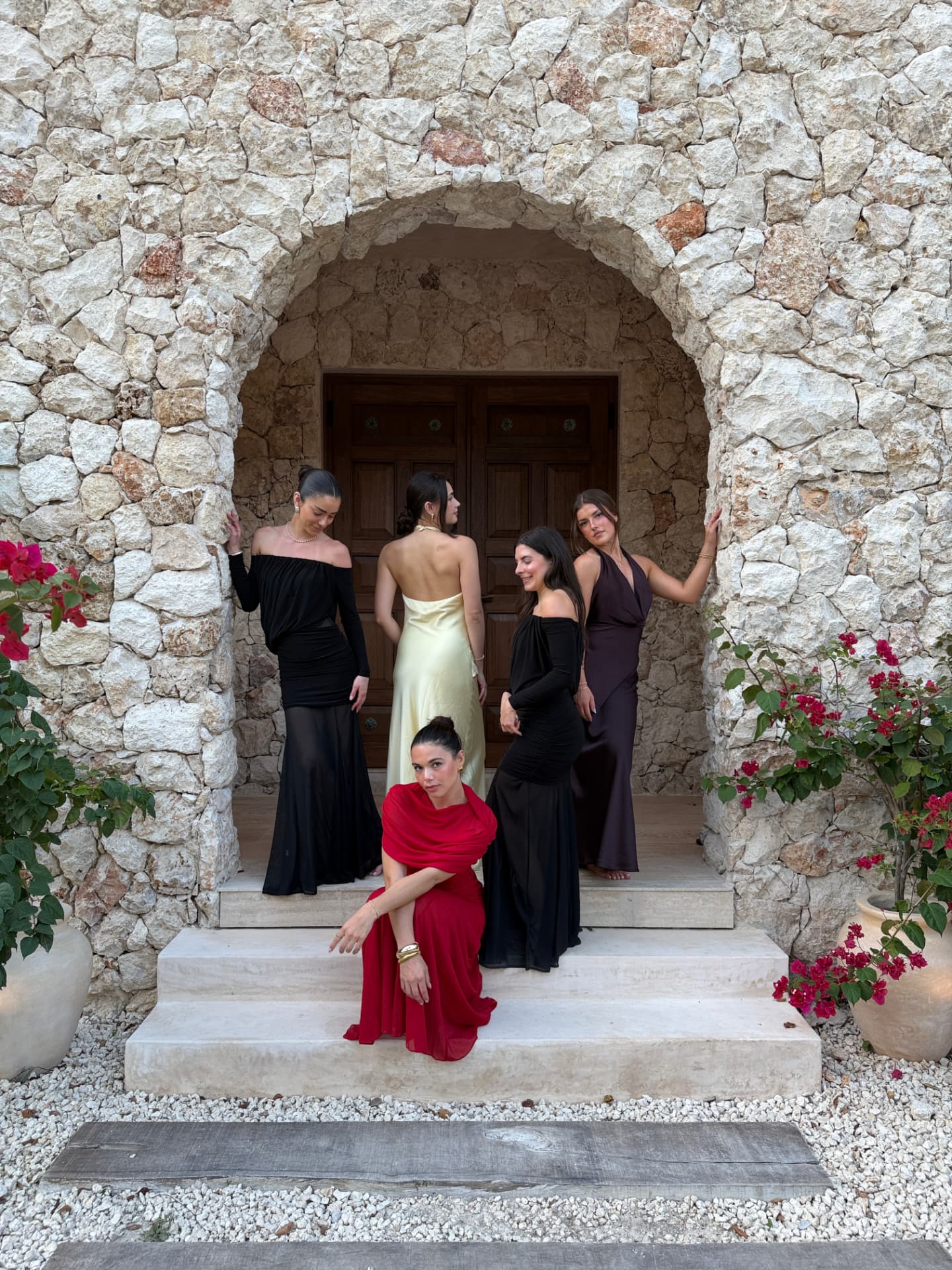 Five women in evening gowns gathered at a stone archway